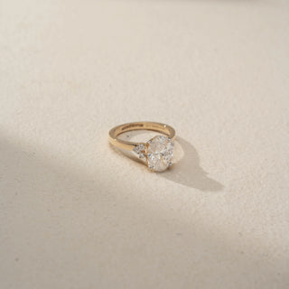 Side view of an oval three-stone engagement ring in 14 KT Yellow Gold, featuring an oval-cut lab-grown diamond flanked by pear-shaped side diamonds, displayed against a beige backdrop.
