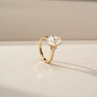 Angled side view of an oval three-stone engagement ring in 14 KT Yellow Gold, featuring an oval-cut lab-grown diamond flanked by pear-shaped side diamonds, displayed against a beige backdrop.