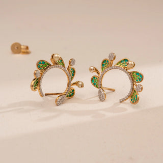 Top-down view of Reia’s 14 KT Yellow Gold Emerald Enamel Winged Diamond Earrings. It highlights the intricate green enamel feather patterns and round brilliant diamonds against a cream backdrop.