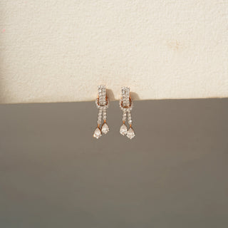Front view of diamond drop earrings in rose gold, featuring pavé-set diamond accents and dangling pear-shaped diamonds. Displayed against a neutral backdrop, the elegant design offers graceful sparkle.