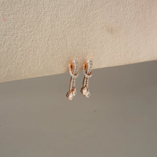 Side view of rose gold diamond drop hoop earrings featuring pavé-set hoops, diamond-accented links, and pear-shaped dangling stones against a neutral textured backdrop.