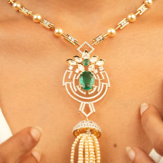 The view of a model styling a female necklace of size 22 secured with a spring lock showcasing a 14 KT yellow gold pearl necklace having a lab-grown white pearl chain along with a pendant featuring an intricate design made out of green Emerald and diamonds with a total of 3.79 carats having a sparkling green Emerald at its centre suspended by a tassel of pearls weighing 24.3 grams for an elegant vintage look perfect for a special occasion.