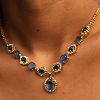 The view of a model styling a female necklace of size 16 showcasing a 14 KT yellow gold necklace secured with a spring lock featuring a symmetrical pattern of lab-grown blue Sapphires with a total product weight of 29.406 grams in a diamond-studded setting with a total of 2.37 carat finished with a central blue Sapphire hanging at the bottom creating a classy look for any occasion.