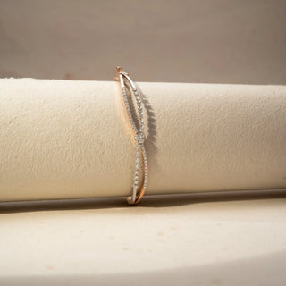 Side view of a diamond bracelet in 14 KT Dual Gold, featuring intertwined pavé-set diamond strands crossing at a floral diamond cluster, displayed against a soft beige backdrop.
