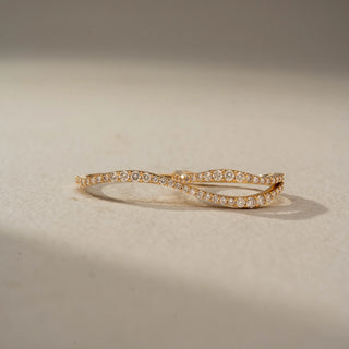 Front view of an infinity diamond band in 14 KT Yellow Gold, featuring intertwined wave-inspired bands adorned with pavé-set round lab-grown diamonds in a graceful crossover design, displayed against a beige backdrop.