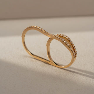 Angled view of an infinity diamond band in 14 KT Yellow Gold, featuring intertwined wave-inspired bands adorned with pavé-set round lab-grown diamonds in a graceful crossover design, displayed against a beige backdrop.