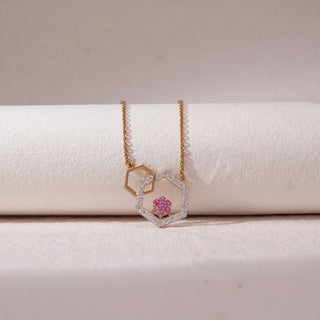 Top view of geometric hexagon pendant necklace in yellow gold, featuring an open hexagon with pavé lab-grown diamonds and a pink stone cluster centre. Placed on a textured beige surface, the design casts a soft shadow.
