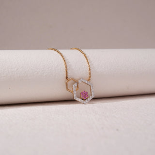 Angled view of geometric hexagon pendant necklace in yellow gold, featuring an open hexagon with pavé lab-grown diamonds and a pink stone cluster centre. Draped over a soft beige form, the design casts a gentle shadow.
