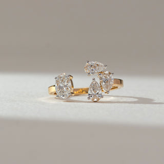 Front view of multi-stone diamond ring in yellow gold, featuring oval and pear-shaped lab-grown diamonds arranged in a clustered setting. Placed on a soft beige surface, the design reflects bold brilliance.
