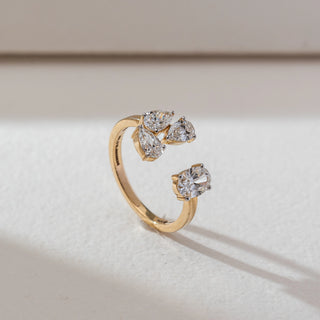 Side view of open cluster diamond ring in yellow gold, featuring round and pear-shaped lab-grown diamonds in a floral arrangement. Placed on a textured beige surface, the design casts a soft elongated shadow.
