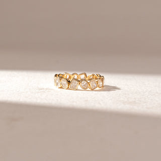Front view of eternity band in yellow gold with mixed-shape diamonds in bezel settings, set on a soft neutral surface, highlighting varied geometry and continuous, radiant sparkle.
