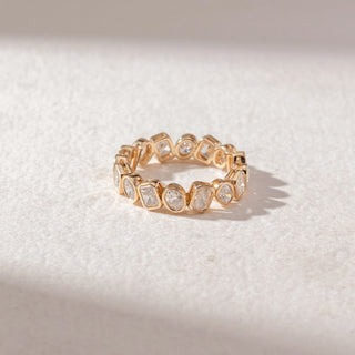 Top view of eternity band in yellow gold with mixed-shape diamonds in bezel settings, set on a soft neutral surface, highlighting varied forms and continuous, refined sparkle.
