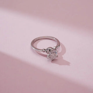 Top view of Reia’s 14 KT White Gold Elegant Silver Knot Solitaire Diamond Ring. It highlights the six-prong solitaire diamond and the intricate knot detail on the band against a pink backdrop.