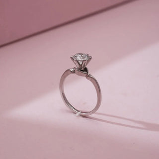 Side view of Reia’s 14 KT White Gold Elegant Silver Knot Solitaire Diamond Ring. It showcases the high six-prong setting and the sleek knot design on the band against a soft pink backdrop.