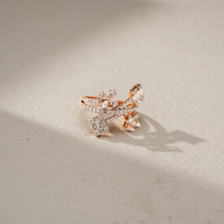 Side view of a floral crossover statement ring in 14 KT Rose Gold, featuring emerald-cut lab-grown diamond clusters, pear-shaped floral motifs, and intertwined pavé diamond bands in an open wrap design, displayed against a beige backdrop.