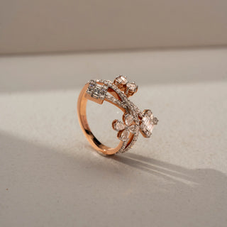 Angled view of a floral crossover statement ring in 14 KT Rose Gold, featuring emerald-cut lab-grown diamond clusters, pear-shaped floral motifs, and intertwined pavé diamond bands in an open wrap design, displayed against a beige backdrop.