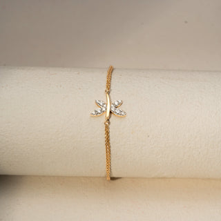 Front view of a dragonfly-inspired diamond bracelet in 14 KT Yellow Gold, featuring marquise-shaped diamond wings on a delicate chain, displayed against a soft beige backdrop.
