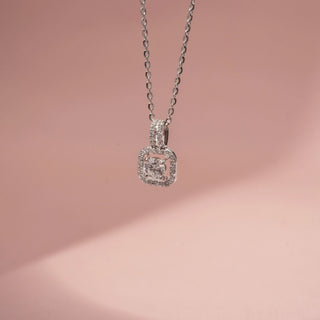 Close up view of a diamond halo pendant necklace in White Gold, featuring a round lab-grown diamond set within a cushion-shaped diamond frame and suspended from a delicate chain against a soft pink backdrop.