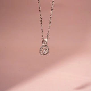 Side view of a diamond halo pendant necklace in White Gold, featuring a round lab-grown diamond within a cushion-shaped diamond frame with elevated settings, suspended from a delicate chain against a soft pink backdrop.