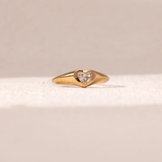 Front view of heart diamond ring in yellow gold, featuring a centred heart-shaped lab-grown diamond set within a smooth sculpted band. Placed on a soft beige surface, the design reflects subtle elegance.
