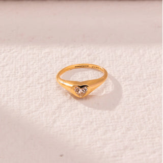Top view of heart diamond ring in yellow gold, featuring a centred heart-shaped lab-grown diamond set within a smooth sculpted band. Placed on a textured beige surface, the design casts a soft shadow.
