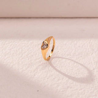Side view of heart diamond ring in yellow gold, featuring a centred heart-shaped lab-grown diamond set within a smooth sculpted band. Placed on a textured beige surface, the design casts a soft shadow.
