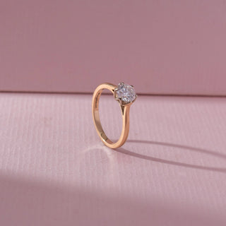 Angled view of round diamond solitaire ring in yellow gold with a slim band, set on a soft pink textured background, highlighting classic elegance and radiant sparkle.
