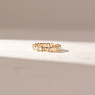 Front view of eternity band in yellow gold with emerald-cut diamonds in bezel settings, set on a soft neutral surface, highlighting uniform geometry and continuous, refined sparkle.

