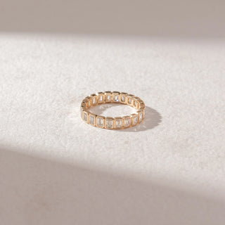 Top view of eternity band in yellow gold with emerald-cut diamonds in bezel settings, set on a soft neutral surface, highlighting continuous design and clean, geometric sparkle.
