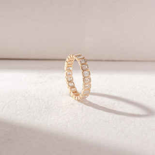 Angled view of eternity band in yellow gold with emerald-cut diamonds in bezel settings, set on a soft neutral background, highlighting structured design and continuous, refined sparkle.
