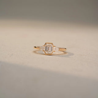 Front view of an emerald-cut three-stone ring in 14 KT Yellow Gold, featuring an emerald-cut lab-grown diamond flanked by pear-shaped side diamonds, displayed against a beige backdrop.