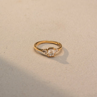Side view of an emerald-cut three-stone ring in 14 KT Yellow Gold, featuring an emerald-cut lab-grown diamond flanked by pear-shaped side diamonds, displayed against a beige backdrop.
