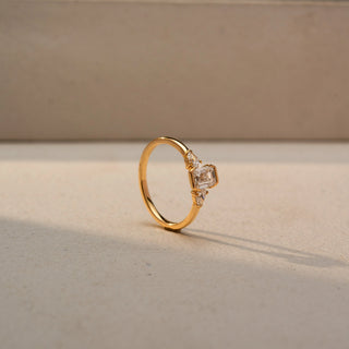 Angled view of an emerald-cut three-stone ring in 14 KT Yellow Gold, featuring an emerald-cut lab-grown diamond flanked by pear-shaped side diamonds, displayed against a beige backdrop.