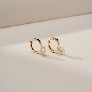 Angled front view of marquise solitaire drop earrings in 14 KT Dual Gold, featuring marquise-cut lab-grown diamonds suspended from sleek huggie hoops, displayed on a beige backdrop.