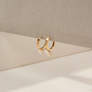 Angled view of yellow gold lab-grown diamond hoop earrings featuring a round brilliant diamond and marquise-shaped diamond drop charms, displayed on a neutral jewellery stand.