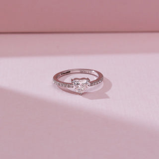 Top view of a 14KT white gold ring featuring a heart-shaped center diamond in a three-prong setting, with a slim pavé-set diamond band, displayed on a soft pink background.