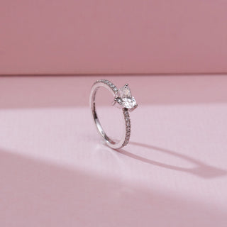 Angled side view of a 14KT white gold ring featuring a heart-shaped center diamond in a three-prong setting, with a slim pavé diamond band, displayed on a soft pink background.