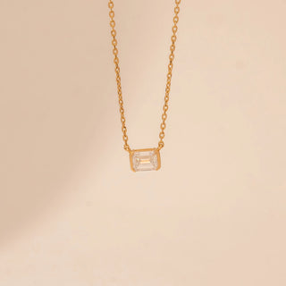 Close-up view of a 14KT yellow gold necklace, featuring a rectangular emerald-cut diamond pendant. Set against a beige backdrop.