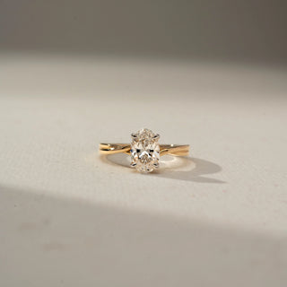 Front view of an oval-cut solitaire diamond ring featuring a twisted yellow gold band and four-prong setting, showcased on a neutral surface in soft natural light.