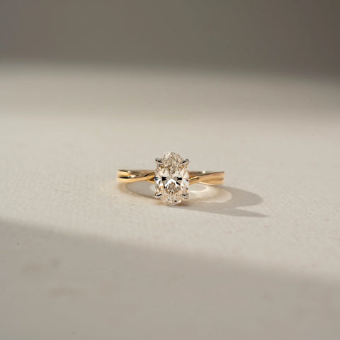 Front view of an oval-cut solitaire diamond ring featuring a twisted yellow gold band and four-prong setting, showcased on a neutral surface in soft natural light.