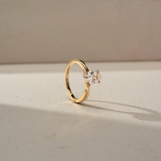 Angled view of a yellow gold solitaire ring featuring a round brilliant-cut diamond in a four-prong setting, displayed on a neutral surface with soft natural lighting.