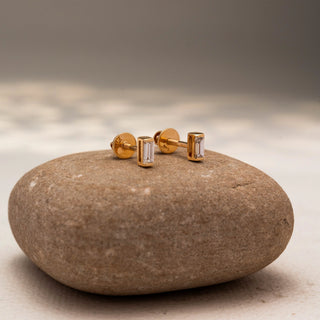Side view of baguette diamond stud earrings in 14 KT Yellow Gold, featuring sleek bezel-set rectangular diamonds, displayed on a textured stone surface.