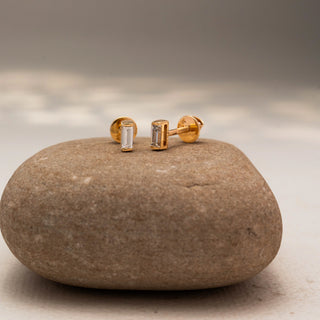 Angled side view of baguette diamond stud earrings in 14 KT Yellow Gold, featuring sleek bezel-set rectangular diamonds, displayed on a textured stone surface.