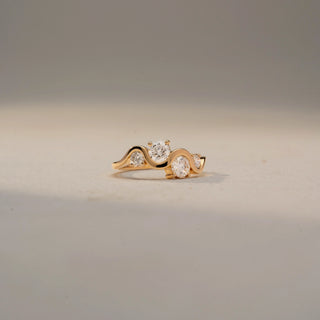 Front view of a three-stone wave ring in 14 KT Yellow Gold, featuring round and oval lab-grown diamonds nestled within a flowing curved band, displayed against a beige backdrop.