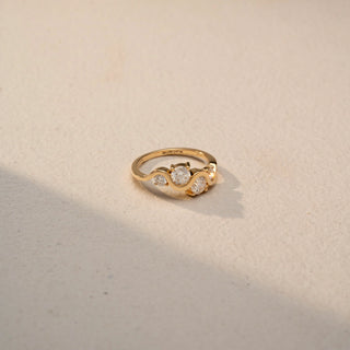 Top view of a three-stone wave ring in 14 KT Yellow Gold, featuring round and oval lab-grown diamonds nestled within a flowing curved band, displayed against a beige backdrop.