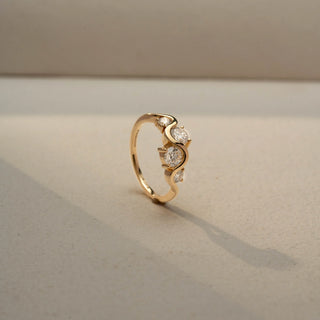 Angle view of a three-stone wave ring in 14 KT Yellow Gold, featuring round and oval lab-grown diamonds nestled within a flowing curved band, displayed against a beige backdrop.