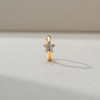 Front view of a delicate gold hoop earring featuring a floral diamond cluster charm, displayed on a soft neutral surface with warm natural light and a subtle shadow.