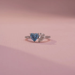 Front view of a toi et moi ring in White Gold, featuring blue and white heart-shaped gemstones set side by side on a pavé-set diamond band, displayed against a soft pink backdrop.