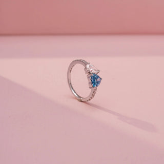 Angled view of a toi et moi ring in White Gold, featuring blue and white heart-shaped gemstones set side by side on a pavé-set diamond band, displayed against a soft pink backdrop.