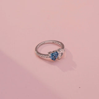 Side view of a toi et moi ring in White Gold, featuring blue and white heart-shaped gemstones set side by side on a pavé-set diamond band, displayed against a soft pink backdrop.